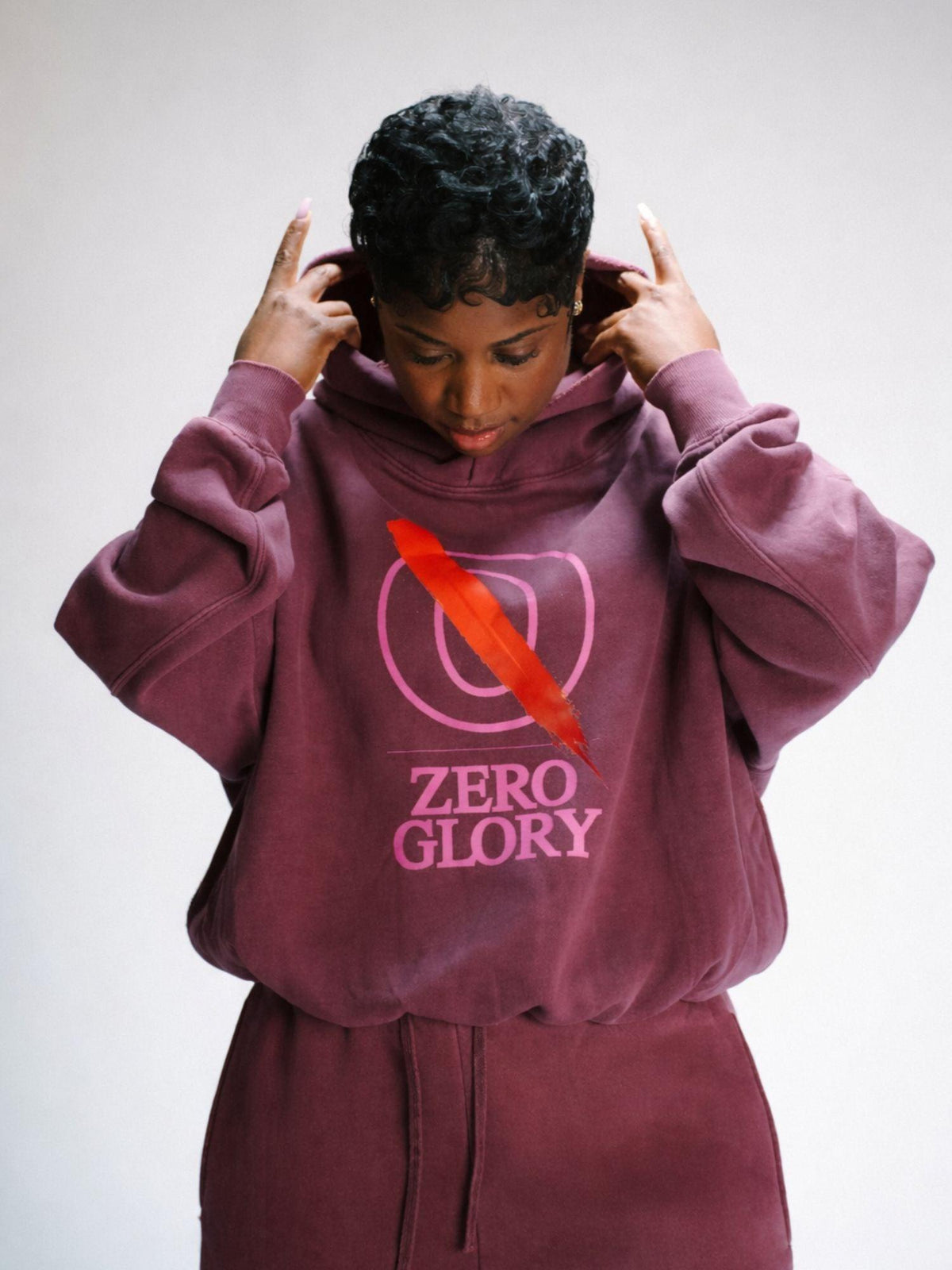 Fall 2026 signature set — Woman wearing maroon hoodie with 'Zero Glory' graphic and red feather design, adjusting hood