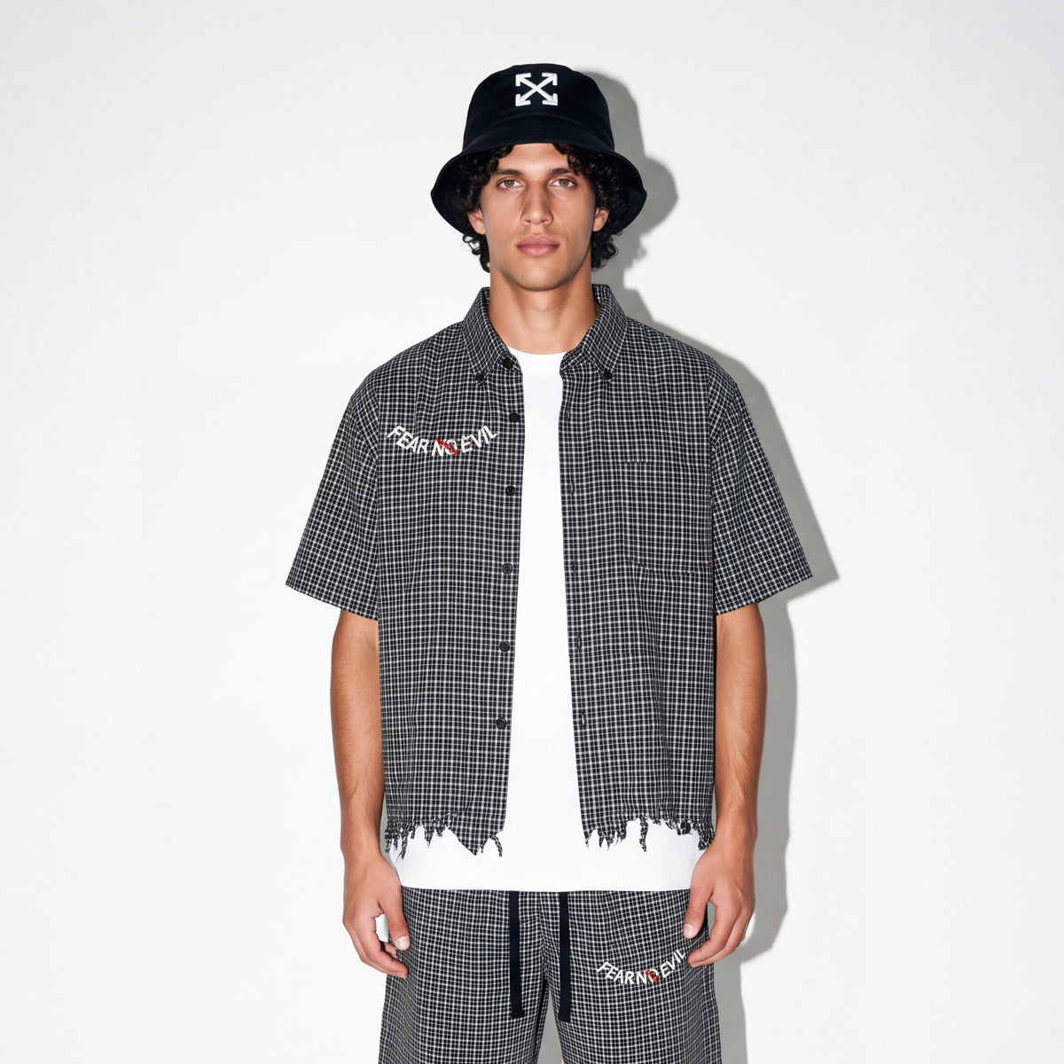 A male model stands against a white studio background wearing a matching black and white gingham outfit consisting of a short-sleeve button-up shirt with raw, frayed hems, matching shorts, a white t-shirt underneath, black crew socks, and black-and-white sneakers. He is also wearing a black bucket hat with a white skull graphic.