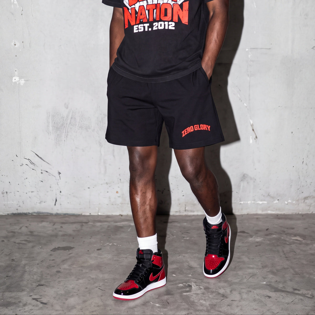 A man stands against a concrete wall wearing a black graphic t-shirt and matching black shorts with red and white text, paired with black and red sneakers.