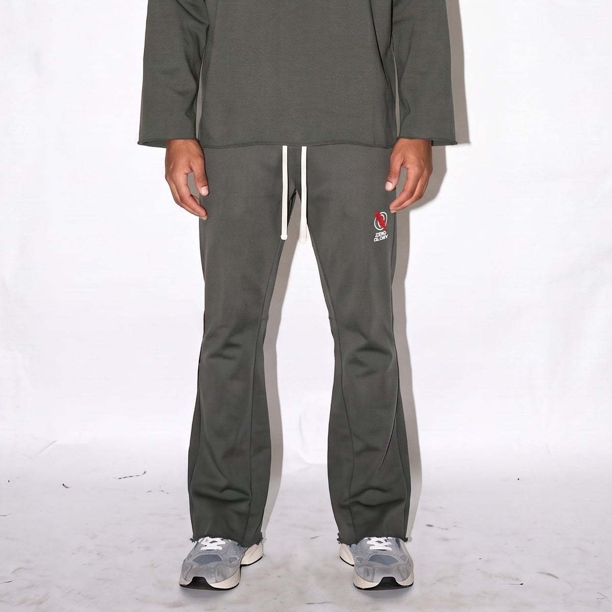 A man stands against a white studio backdrop wearing a dark gray long-sleeve sweatshirt and matching dark gray sweatpants with a small red and white logo on the chest and thigh.