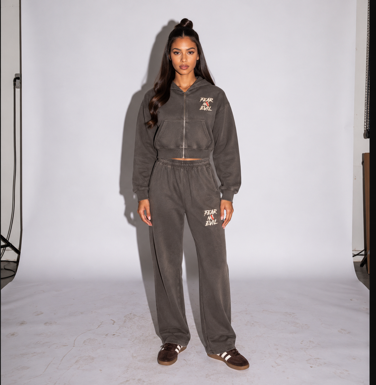 A woman stands in a studio wearing a matching dark gray zip-up hoodie and sweatpants set with small white text graphics on the chest and thigh, paired with brown shoes.