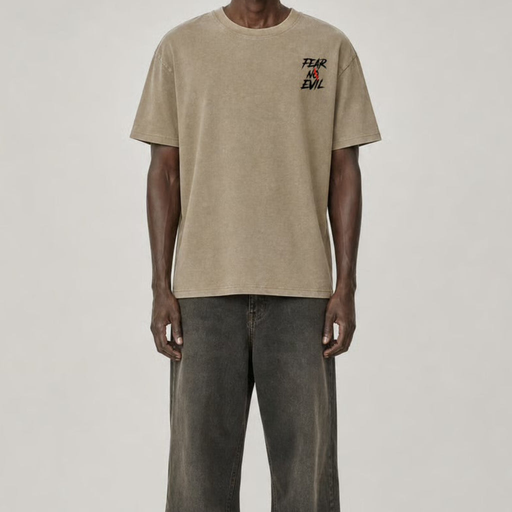 A male model stands against a light gray studio background wearing a beige short-sleeve t-shirt with a small multicolor graphic on the left chest, paired with dark gray baggy jeans and black shoes.