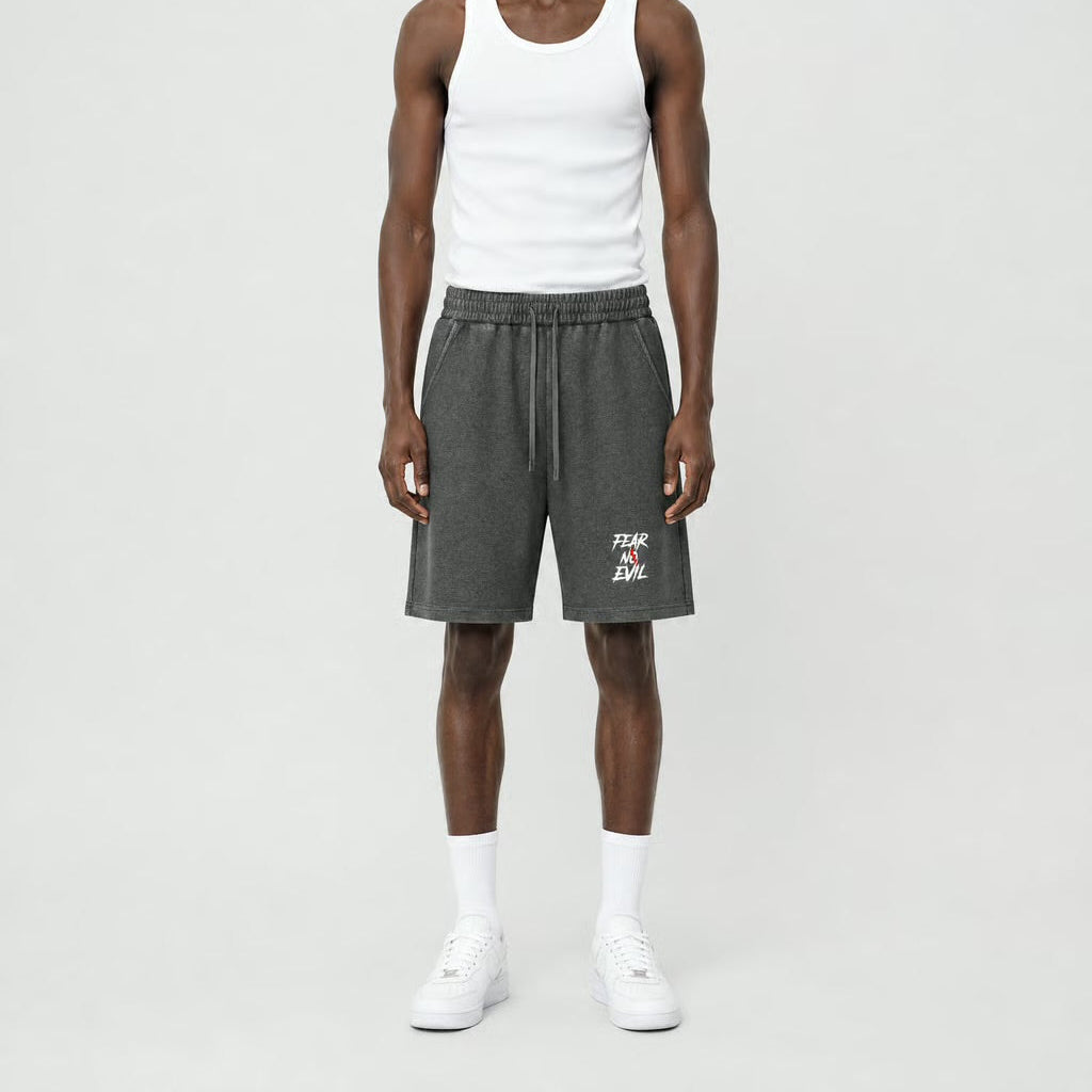 A male model stands against a light gray studio background wearing a white ribbed tank top, dark gray heathered shorts with a small white graphic on the lower left leg, white socks, and white sneakers.
