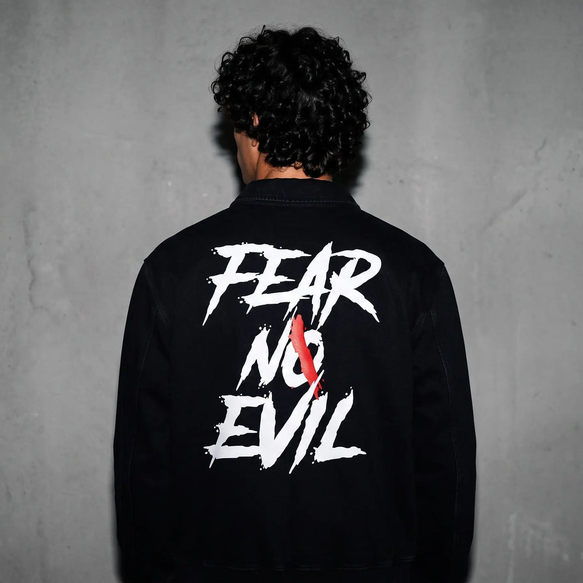 A person viewed from behind wearing a black jacket with a large white graphic reading 'FEAR NO EVIL' and a red ribbon detail on the back, standing against a textured gray wall.