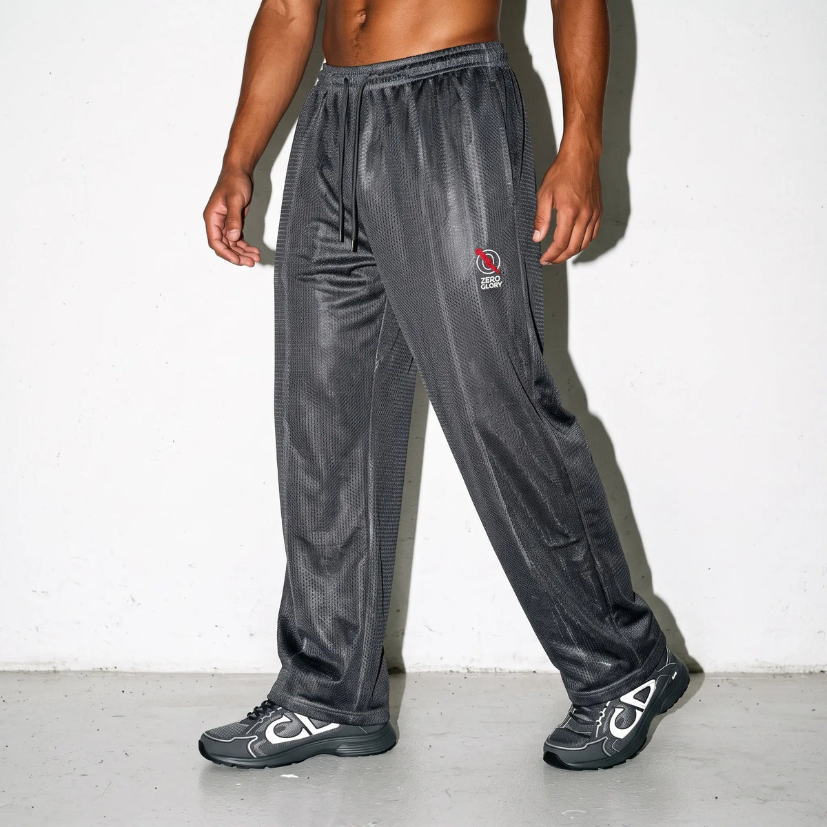 A person modeling dark gray textured athletic pants with a small red and white logo on the left thigh, paired with black and white sneakers, standing against a white wall.