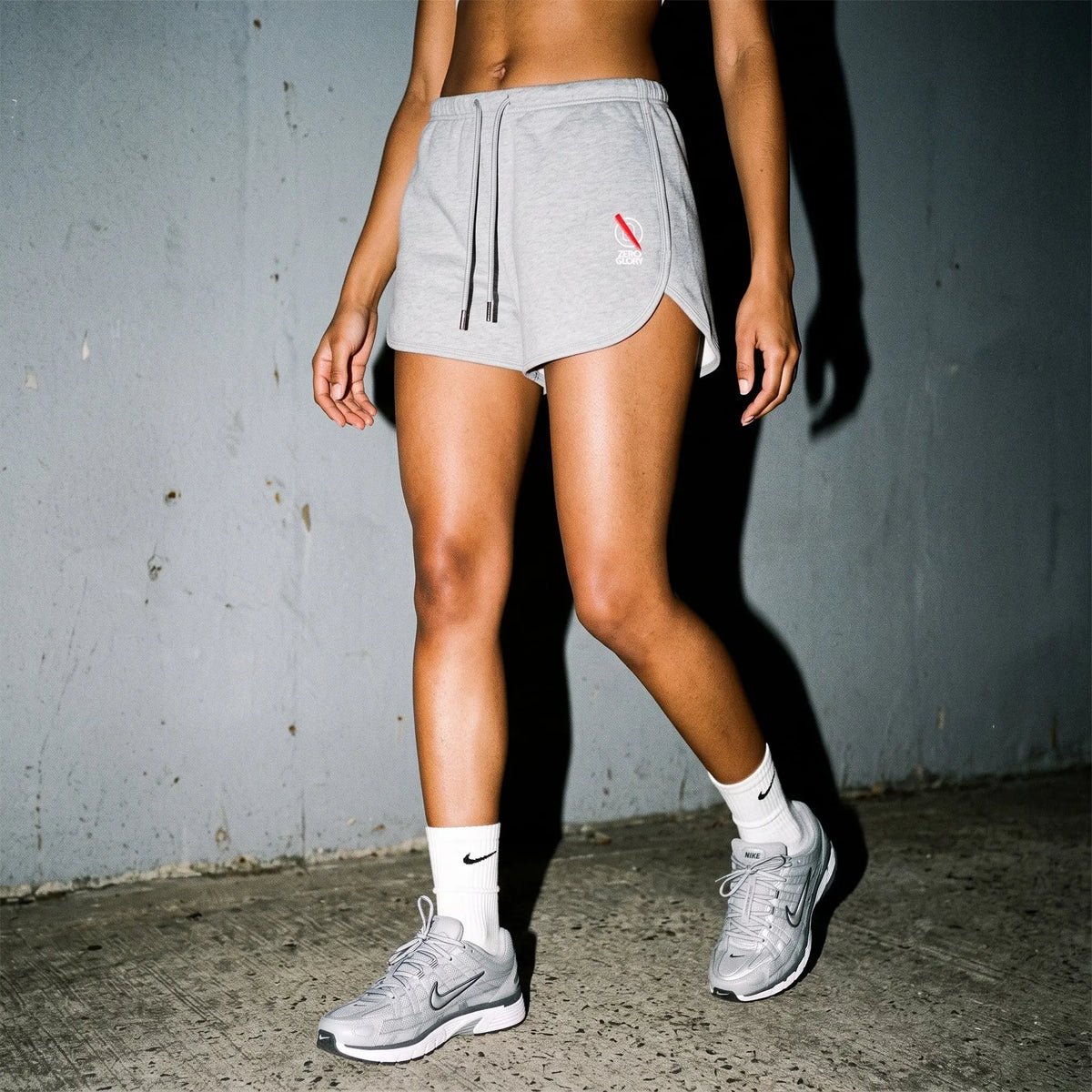 A person wearing light gray athletic shorts, white socks, and gray running shoes, standing against a textured gray wall.