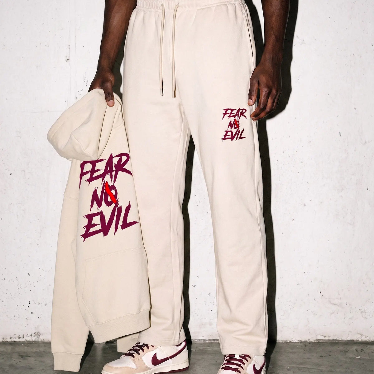 A model wearing beige sweatpants with red 'FEAR AND EVIL' text graphics, holding a matching beige hoodie with the same graphic. The model is also wearing beige and red Nike sneakers, standing against a textured white wall.
