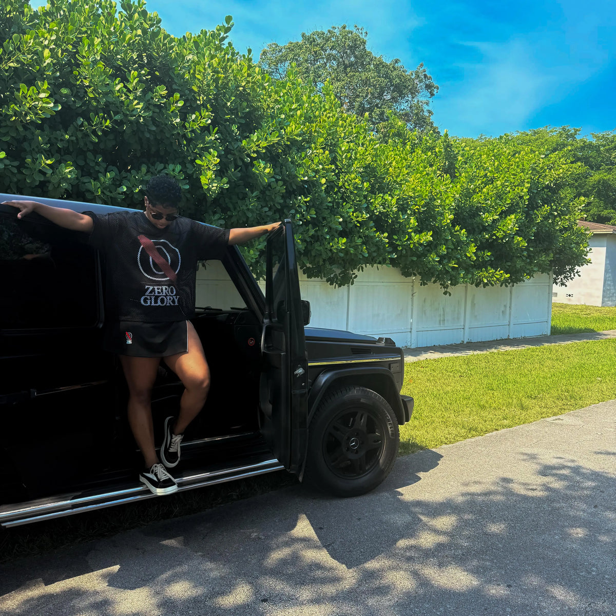 Person in black Christian streetwear for creatives stepping out of a luxury vehicle amidst lush greenery.