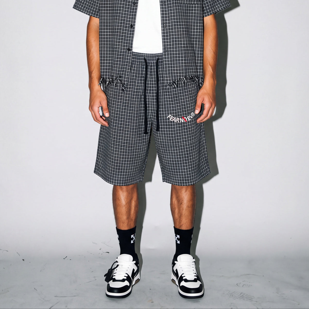 A model stands against a light gray studio backdrop wearing a matching black and white checkered short-sleeve button-up shirt, shorts, and a black bucket hat with a white cross logo. The outfit is styled over a white t-shirt, black socks, and black-and-white sneakers.