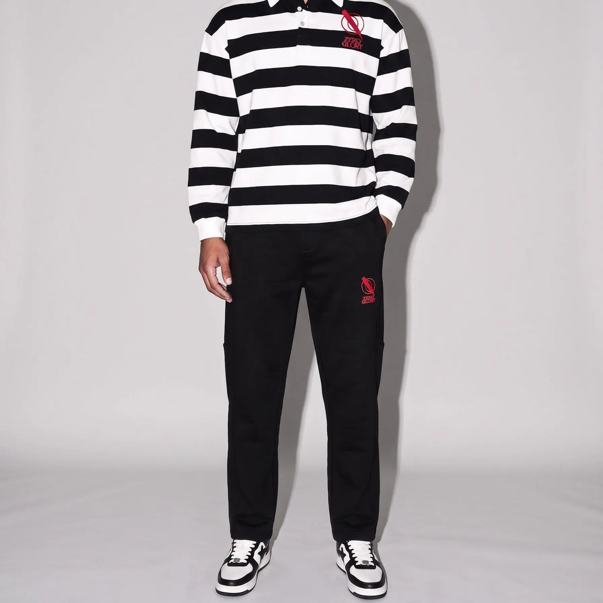 A man modeling a black and white horizontally striped long-sleeve rugby shirt with a red embroidered logo on the chest, paired with black pants and black-and-white sneakers, against a light gray studio background.