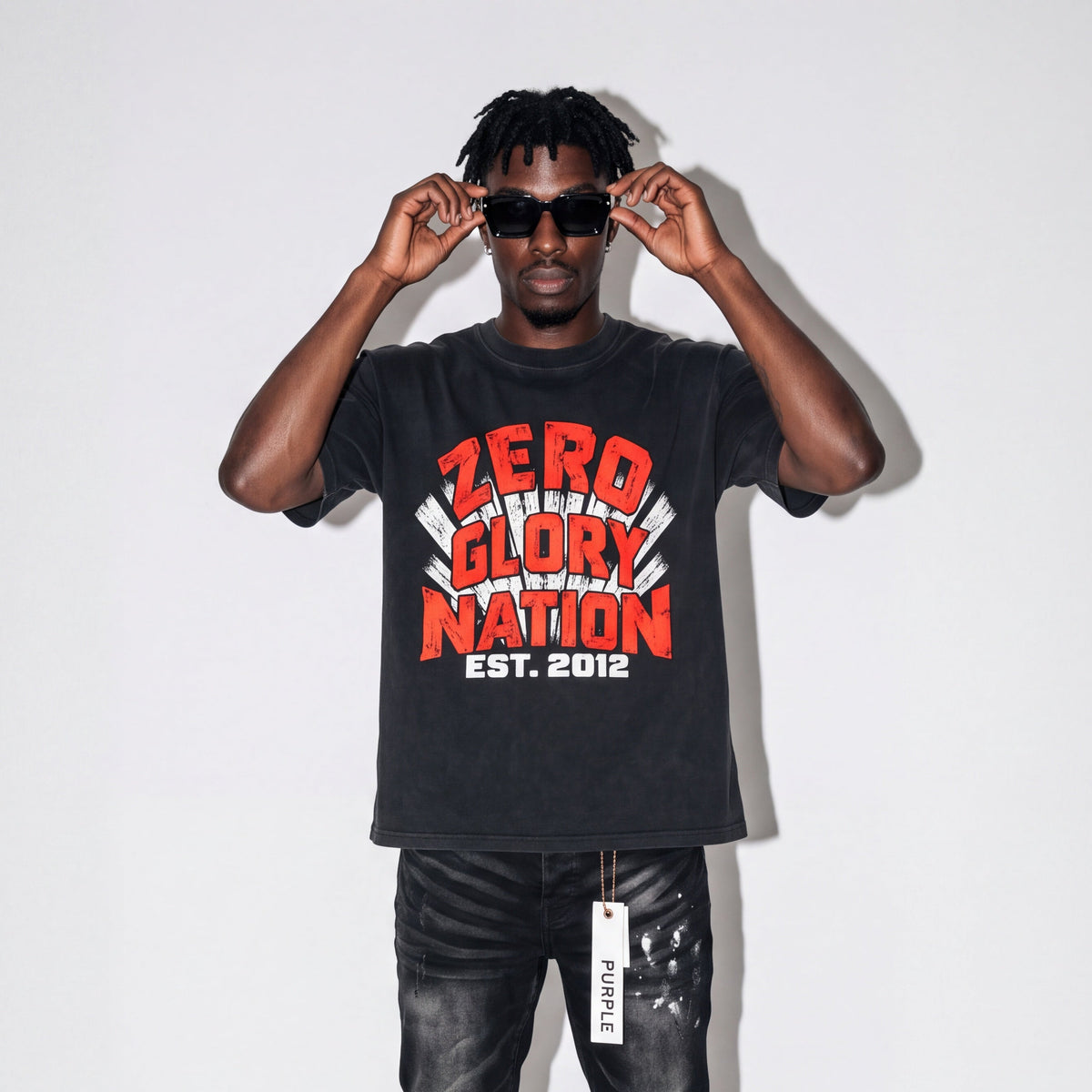 A man modeling a black graphic t-shirt with 'ZERO TOLERANCE GLORY NATION EST. 2012' printed in red and white, paired with black jeans and black/red sneakers, against a white studio background.