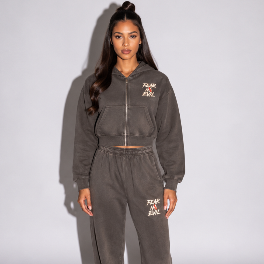 A woman stands in a studio wearing a matching dark gray zip-up hoodie and sweatpants set with small white text graphics on the chest and thigh, paired with brown shoes.