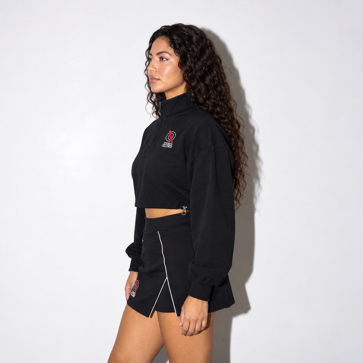 Side view of the Zero Glory Women's Mock-Neck Quarter-Zip Sweatshirt in black worn with the Women's High-Waist Performance Skort