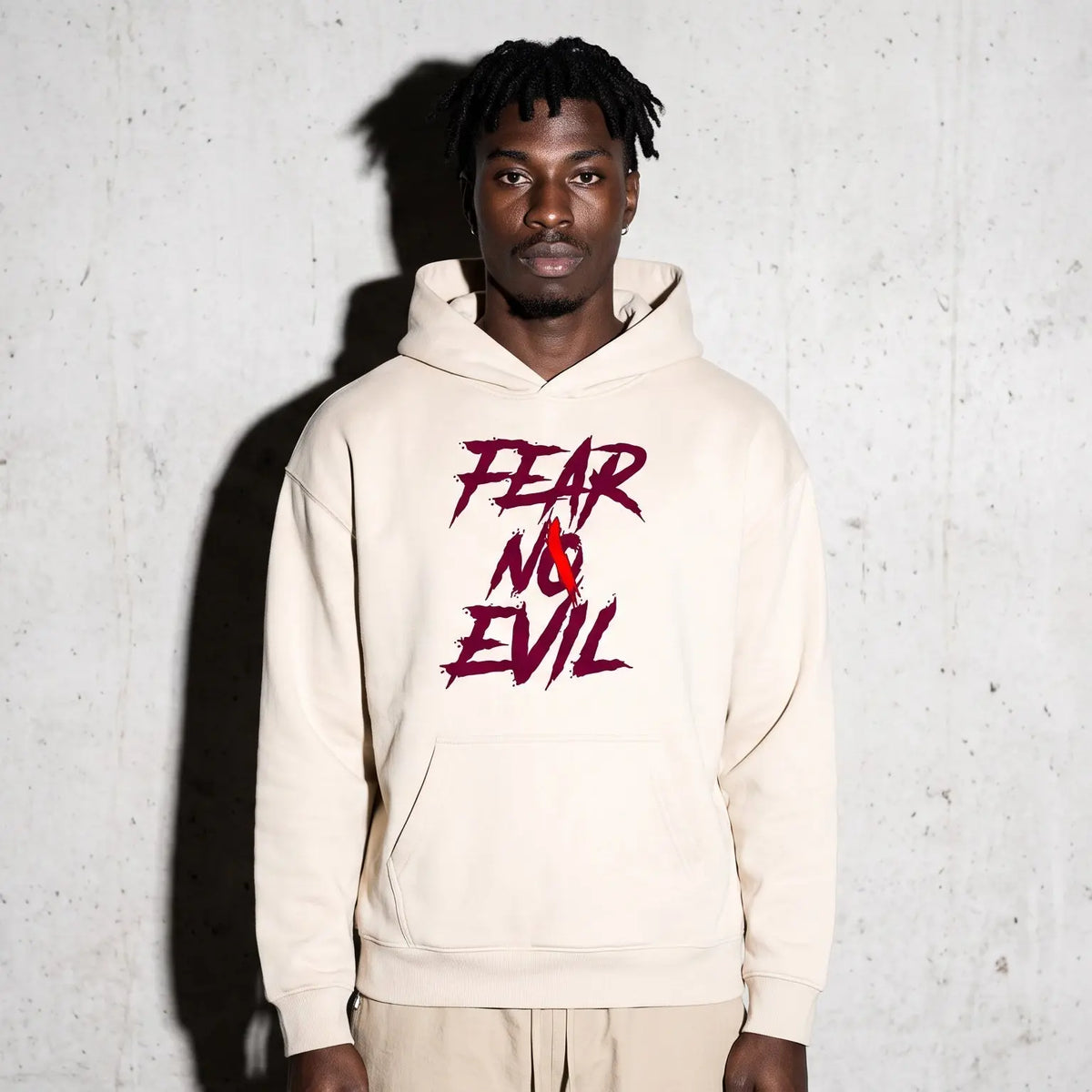 A man wearing a cream-colored hoodie with a red and black 'FEAR NO EVIL' graphic on the chest, standing against a textured concrete wall.