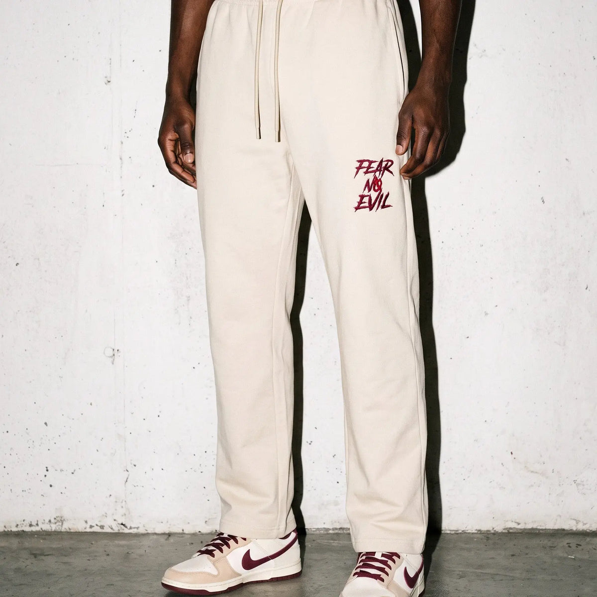 A model wearing off-white sweatpants with a red 'FEAR AND EViL' graphic on the left thigh, paired with matching sneakers, standing against a textured concrete wall.